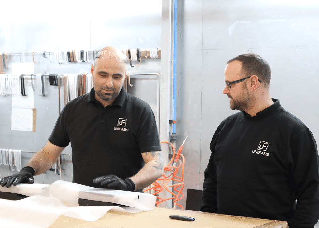 Chris Lawrence Production Manager discusses product packing with sheet metal fabrication team operator at the Unifabs site based in Nuneaton UK. Powder coating line is visible in the background with various coloured hanging hooks.
